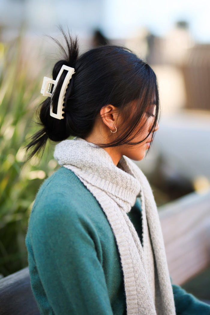 Woman with dark hair tied back with a white hair clip, wearing a green sweater and beige scarf, outdoors.