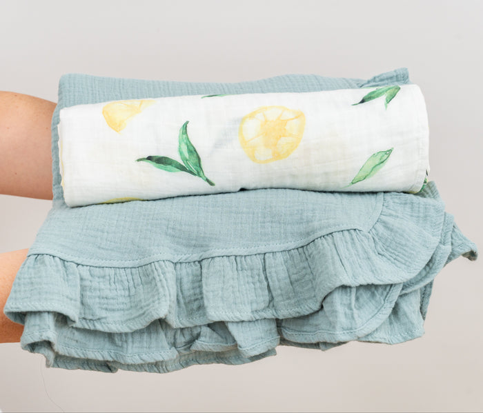 Person holding stacked, folded muslin swaddle with lemon design and green blanket on a light gray background