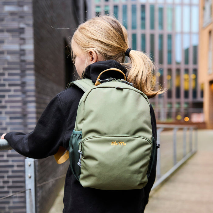 Ela Mo™ Kinderrucksack (3-6 Jahre) | Forrest Green. Mädchen mit einem grünen Rucksack in einem alltäglichen Lifestyle.