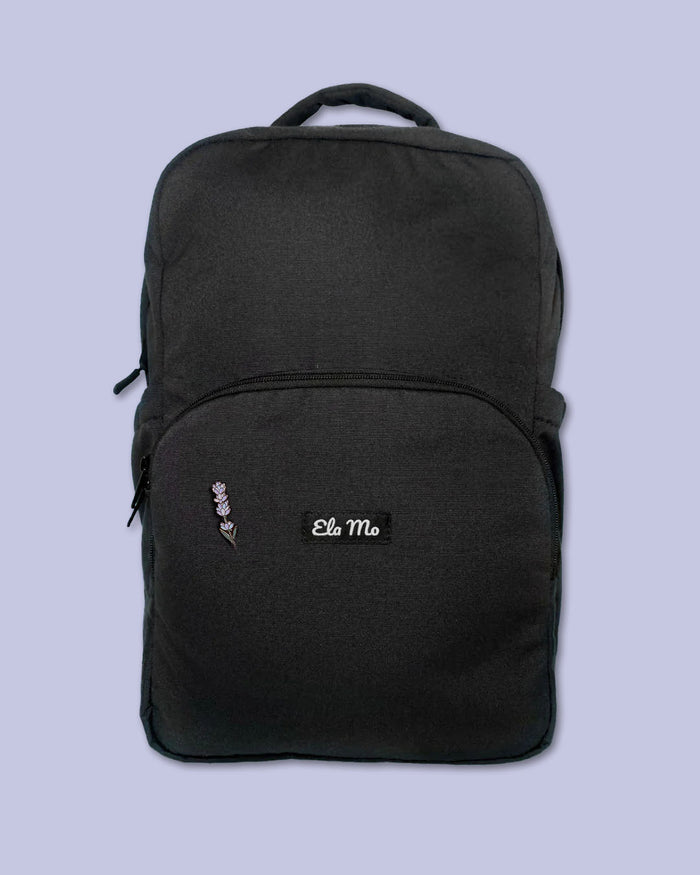 Ela Mo™ Mini Pin für Rucksäcke | Lavender. Ein kleiner Lavendel-Pin am Rucksack, vor lila Hintergrund.