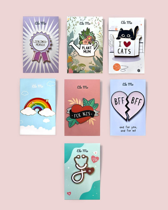 Ela Mo™ Pin für Rucksäcke| Rainbow. Verschiedene Ausführungen von Rucksackpins auf rosa Hintergrund.
