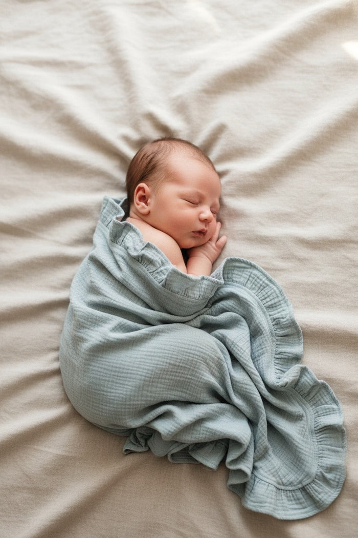 Newborn baby wrapped in a Newborn baby wrapped in a lagoon green heirloom muslin ruffle blanket on a beige surface