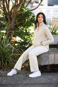 Woman sitting on a wooden bench wearing a beige outfit with white sneakers.