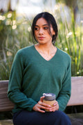 Woman in a green sweater holding a coffee cup outdoors with greenery in the background