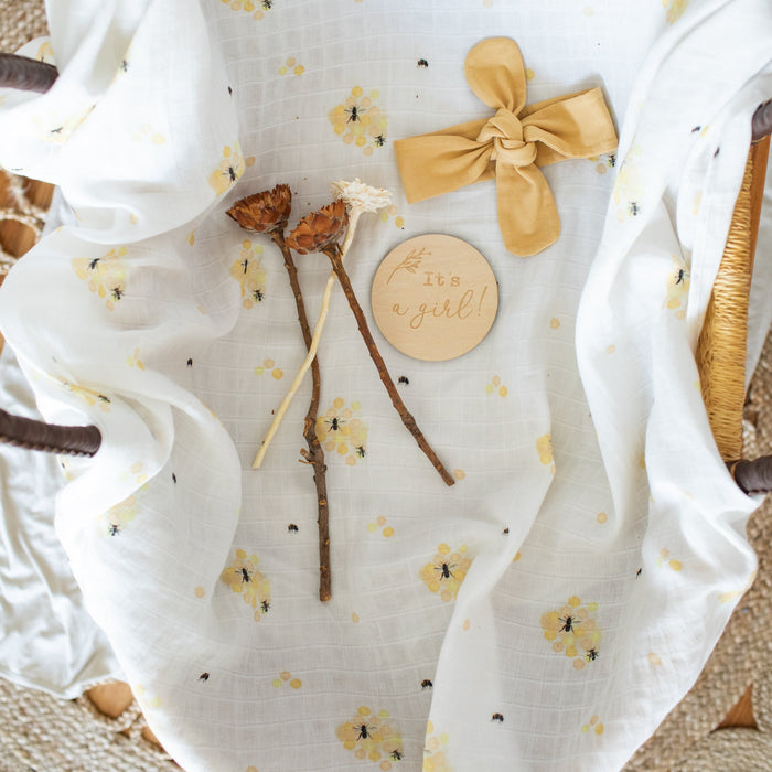 Gender reveal milestone card 'It's a girl' with headband and honey bees organic muslin swaddle.