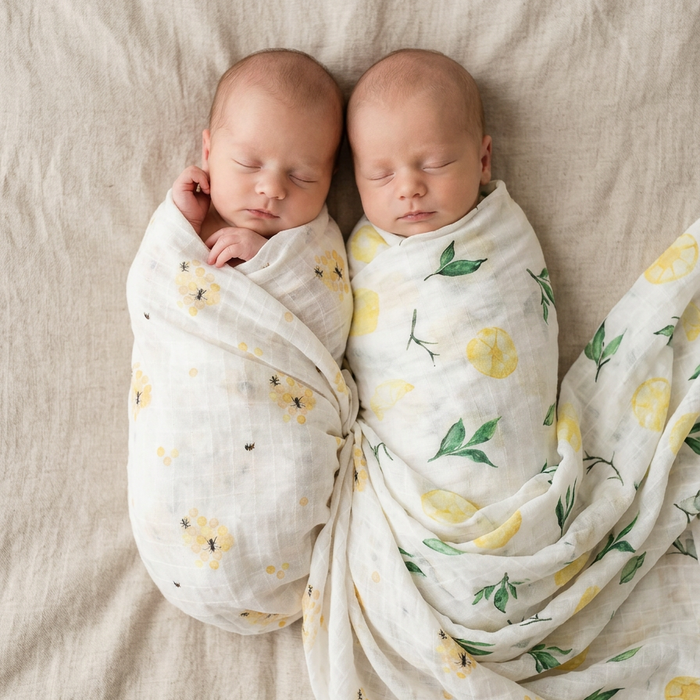 Two newborn babies swaddled in floral-patterned blankets on a beige surface.