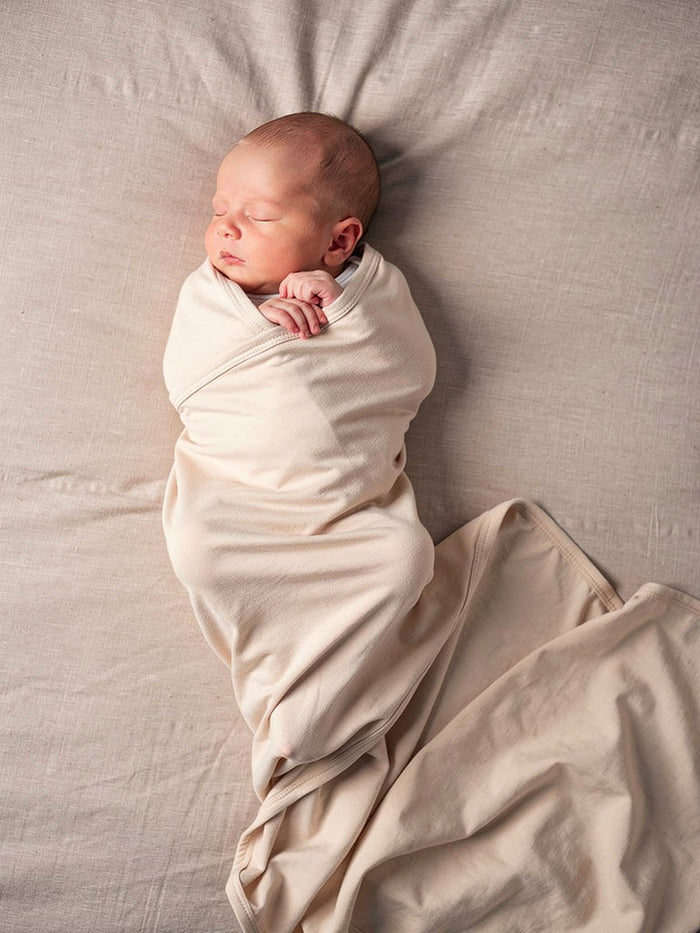 Newborn baby wrapped in an organic jersey swaddle with matching beanie in Fog, creating a calm and soothing newborn moment.