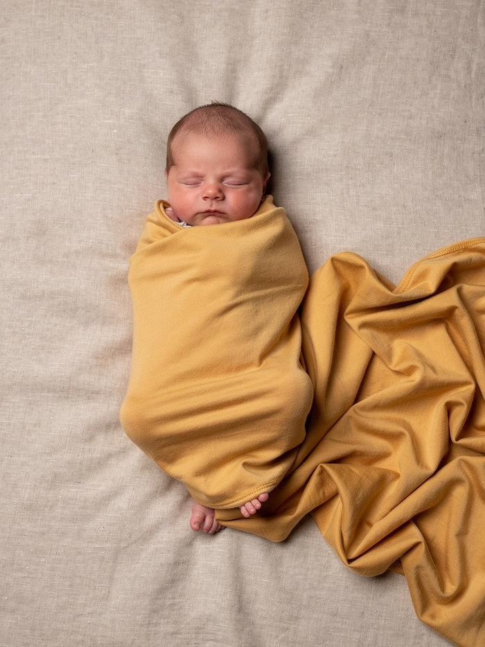 Newborn baby wrapped in an organic jersey swaddle with matching beanie in Turmeric, creating a warm and comforting newborn moment.