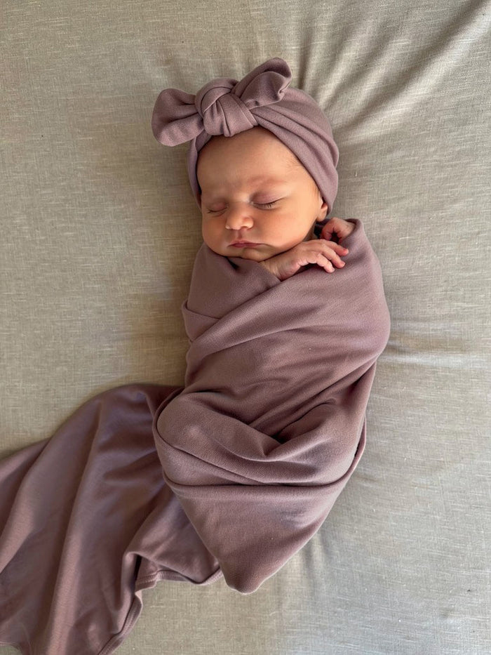 Newborn baby wrapped in an organic jersey swaddle with matching topknot in Amethyst, creating a calm and comforting newborn moment.