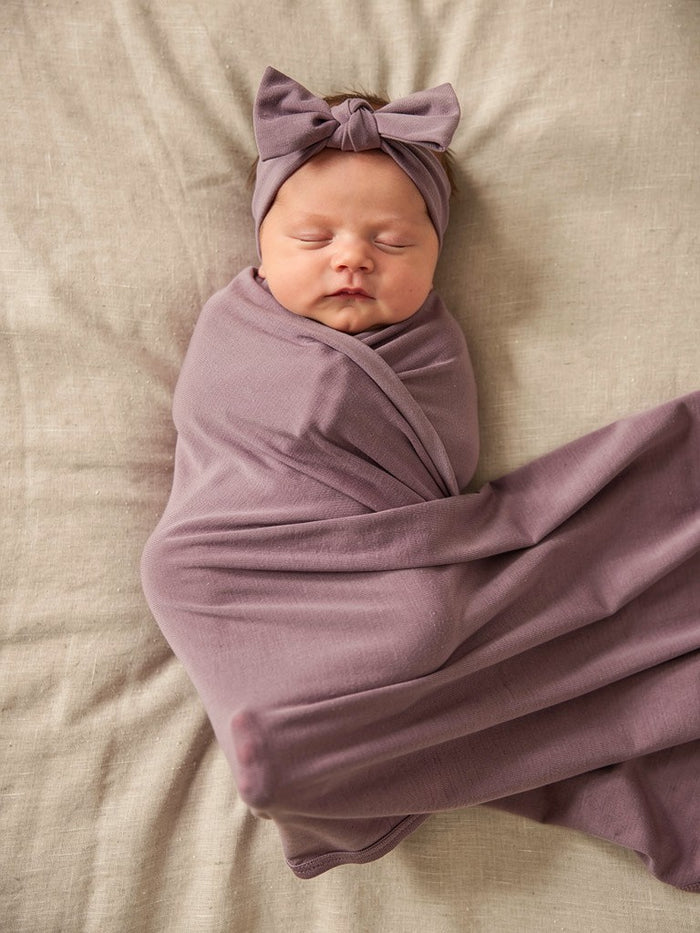 Newborn baby swaddled in a purple jersey wrap with a matching headband on a beige surface