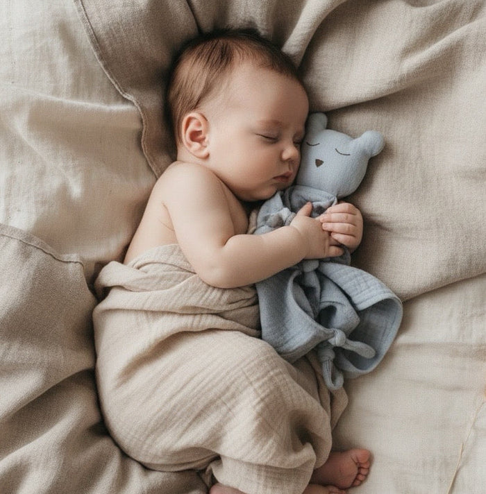 Baby sleeping with a blue muslin bear comforter soft toy on beige bedding