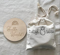 Milestone cards with keepsake pouch. Round wooden disc with rainbow and  'Hello World!' text on a light gray fabric background.