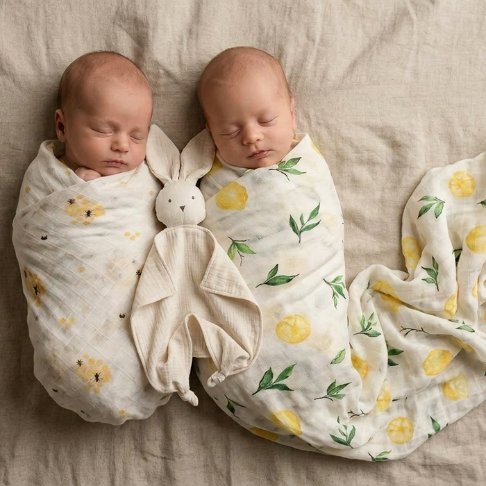 Two babies wrapped in muslin swaddles with lemon pattern and bee pattern a beige blanket.