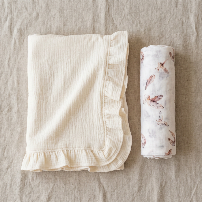 Folded white blanket with ruffled edge and owl printed swaddle on a beige surface